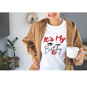 May include: White t-shirt with the text "It's My Birthday" in red and black script, featuring a winking eye and red lips graphic. The person is wearing a brown jacket and holding a white mug.