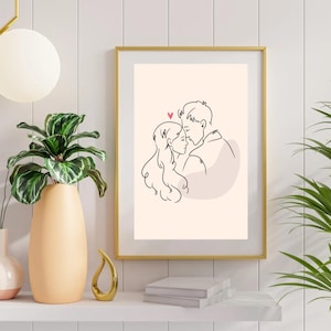 May include: A framed art print featuring a minimalist line drawing of a couple embracing, with a small pink heart above their heads. The artwork is set against a cream background and framed in gold. The print is displayed on a white shelf with decorative items.