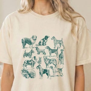 Peut inclure: T-shirt crème avec des illustrations de chiens de couleur sarcelle. Le motif présente diverses races de chiens dans différentes poses, créant une esthétique vintage. Le t-shirt est en matière douce.