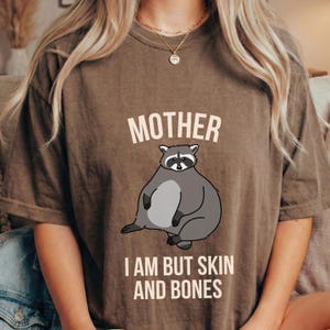 Puede incluir: Camiseta marrón con la palabra "MOTHER" encima de una ilustración de mapache de dibujos animados. Debajo del mapache, el texto dice "I AM BUT SKIN AND BONES". El mapache es gris y blanco.