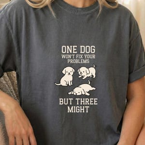May include: Dark grey t-shirt with the text "ONE DOG WON'T FIX YOUR PROBLEMS BUT THREE MIGHT." The shirt features three cartoon puppies in white. One puppy is sitting, one is lying down, and one is on its side.
