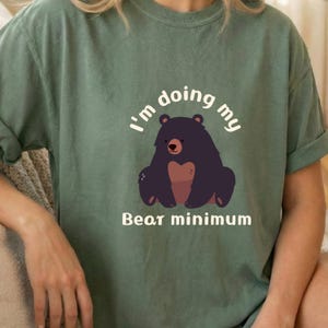 Puede incluir: Camiseta verde con la frase "I'm doing my bear minimum" y una ilustración de oso de dibujos animados. El oso es de color púrpura oscuro con la barriga marrón y la nariz roja. El texto es blanco.