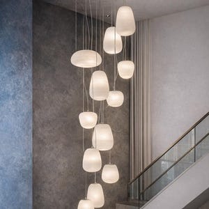 Peut inclure: Un lustre moderne avec plusieurs lampes suspendues blanches et texturées de formes et de tailles variées. Les lumières sont suspendues à un luminaire de plafond circulaire. L'arrière-plan présente un escalier et un mur gris texturé.