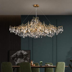 Op de afbeelding: Een goudkleurige kroonluchter met een takachtig ontwerp, versierd met talrijke kristallen hangers. De armatuur hangt boven een eettafel met groene stoelen, tegen een donkerblauwe muur. Het ingewikkelde ontwerp van de kroonluchter voegt elegantie toe.