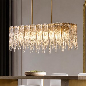 May include: A rectangular chandelier with a gold-coloured frame and multiple clear, icicle-like glass pendants. The light fixture hangs from two gold-coloured rods. The chandelier is suspended above a wooden table.