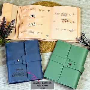 May include: An open, light pink earring display book with various earrings. Two closed leatherette jewelry cases, one blue and one green, are below. The blue case has the name "Sophia" and a floral design. The text "CUSTOM PATTERN AND NAME HERE" is also visible.