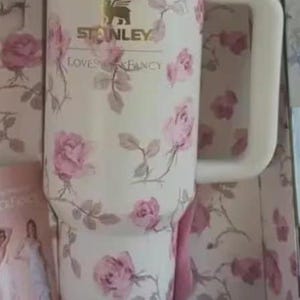May include: A white Stanley tumbler with a pink rose floral design. The tumbler features a large handle and a lid with a straw. The text "STANLEY LOVES FANCY" is printed on the tumbler. The tumbler is presented in a box with a matching floral pattern.