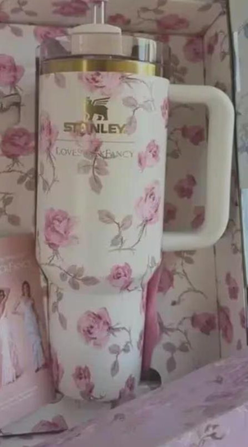 May include: A white Stanley tumbler with a pink rose floral design. The tumbler features a large handle and a lid with a straw. The text "STANLEY LOVES FANCY" is printed on the tumbler. The tumbler is presented in a box with a matching floral pattern.