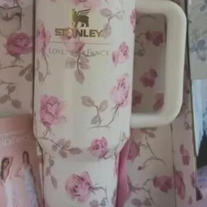 May include: A white Stanley tumbler with a pink rose floral pattern. The tumbler has a large handle and a gold rim. The text "Stanley Loves & Fancy" is visible. It is presented in a pink box.
