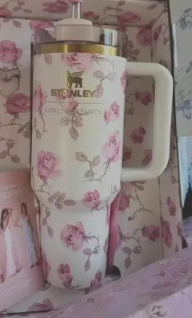 May include: A white Stanley tumbler with a pink rose floral pattern. The tumbler has a large handle and a gold rim. The text "Stanley Loves & Fancy" is visible. It is presented in a pink box.