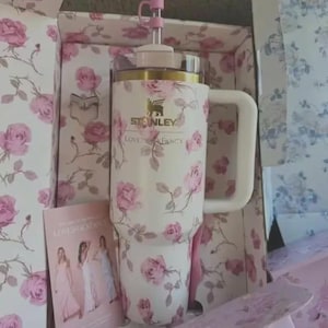 May include: A white Stanley tumbler with a floral rose pattern, a white handle, and a pink bow straw topper. The tumbler is in a matching floral box with the text "LOVES FANCY". A card with images of women in dresses is also visible.