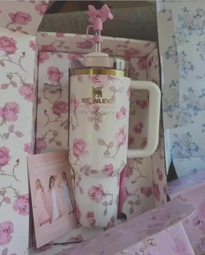 May include: A white Stanley tumbler with a floral rose pattern, a white handle, and a pink bow straw topper. The tumbler is in a matching floral box with the text "LOVES FANCY". A card with images of women in dresses is also visible.