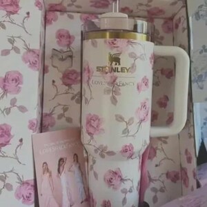 May include: A white Stanley travel mug featuring a pink rose print, a white handle, and a pink straw. The mug is presented in a box with a coordinating floral design. The text "STANLEY" and "LOVESHACKFANCY" are visible.