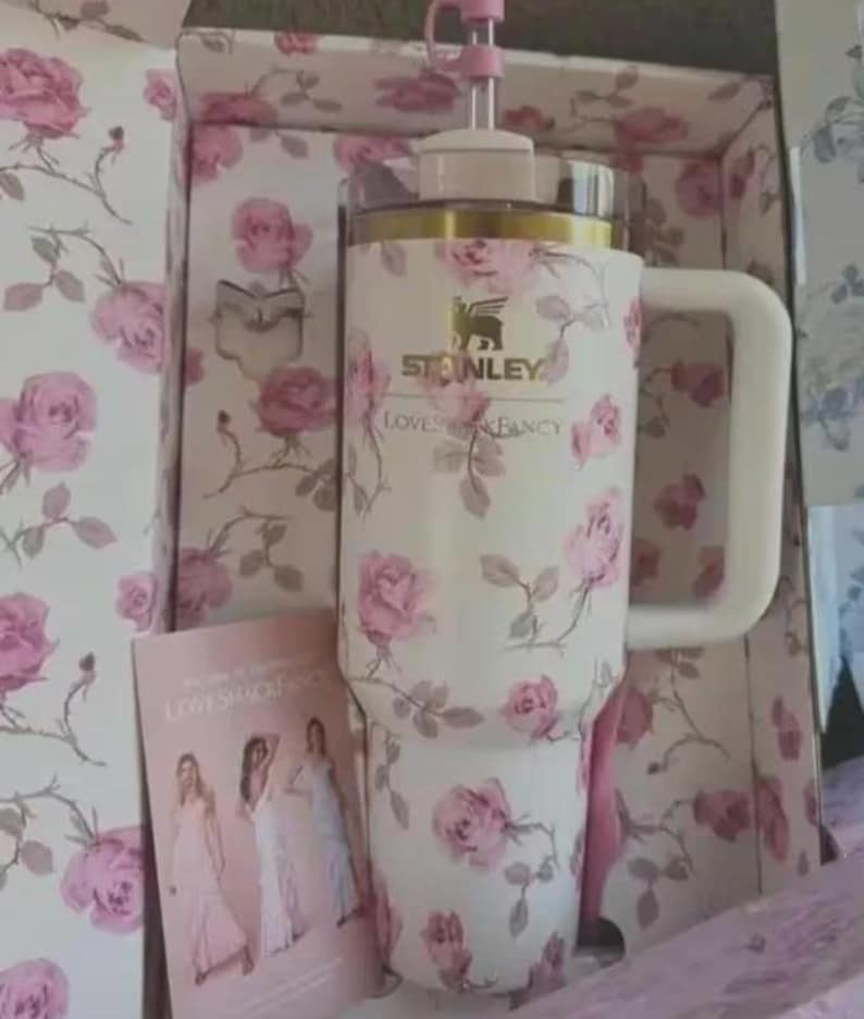 May include: A white Stanley travel mug featuring a pink rose print, a white handle, and a pink straw. The mug is presented in a box with a coordinating floral design. The text "STANLEY" and "LOVESHACKFANCY" are visible.