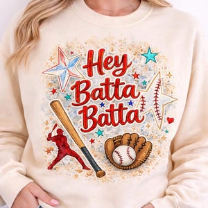 May include: Cream-colored sweatshirt with a baseball-themed graphic. The design features the text "Hey Batta Batta" in red, along with stars, a baseball bat, a baseball glove, and a baseball. The graphic is in a distressed style.
