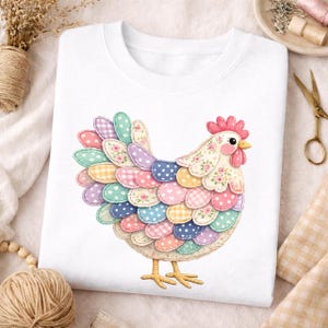 May include: White sweatshirt featuring a colorful, patchwork-style chicken design. The chicken has a red comb, pink cheeks, and a yellow beak. Its feathers are a mix of pastel colors and patterns, including polka dots and floral prints. The design is centered on the front of the shirt.