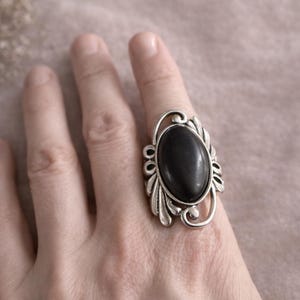 May include: Silver-toned ring featuring an oval, black stone. The ring has an ornate, decorative design around the stone, with a floral or leaf-like pattern. The ring is on a person's finger.