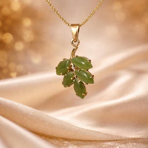 May include: A gold-toned necklace with a pendant featuring a cluster of green, oval-shaped gemstones. The gemstones are arranged to resemble leaves. The necklace hangs against a soft, golden background.