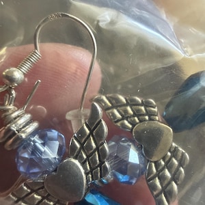 May include: Silver-toned earrings featuring heart and wing charms, accented with faceted blue beads. The earrings have a hook closure and are visible through a clear plastic bag. The metal is stamped with "S925".
