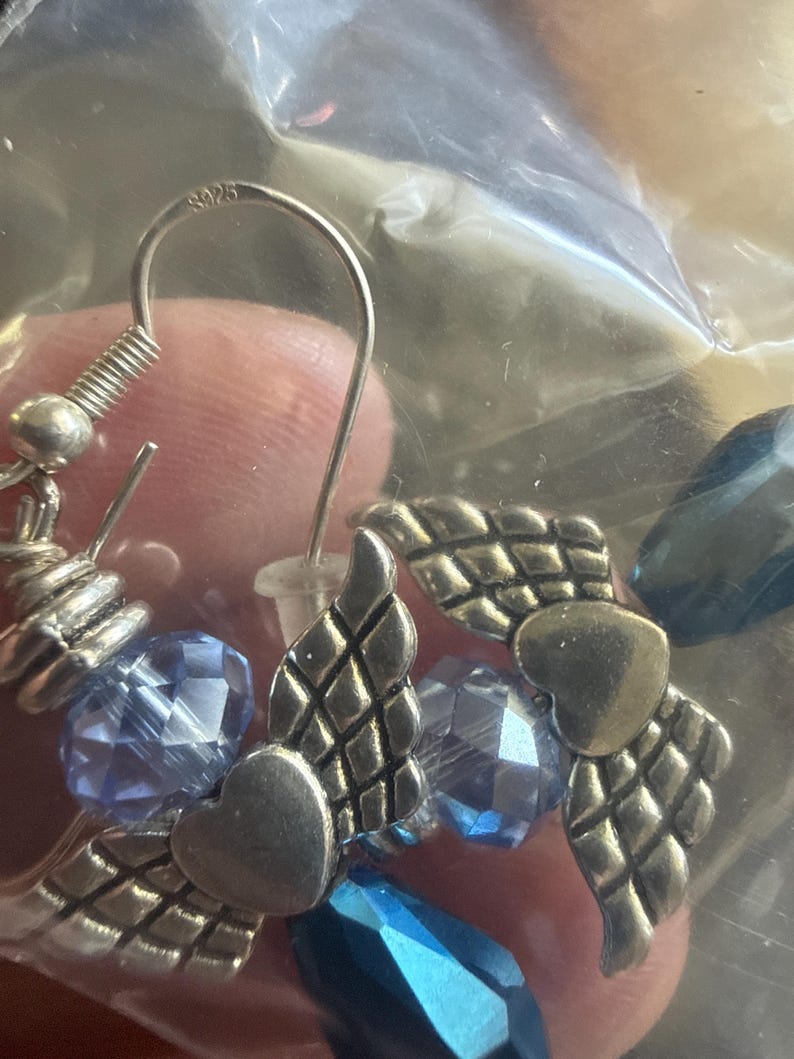 May include: Silver-toned earrings featuring heart and wing charms, accented with faceted blue beads. The earrings have a hook closure and are visible through a clear plastic bag. The metal is stamped with "S925".