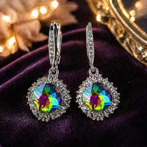 May include: A pair of silver-toned drop earrings. Each earring features a square, iridescent gemstone surrounded by a halo of small, clear crystals. The gemstones display a vibrant array of colors, including green, blue, and purple, against a dark purple background.