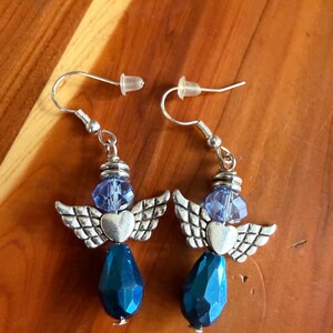 May include: A pair of handmade earrings featuring an angel design. Each earring has a silver-tone heart and wing charm, a blue faceted bead, and a teardrop-shaped blue bead. The earrings hang from silver-tone ear wires.