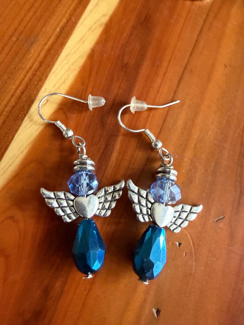 May include: A pair of handmade earrings featuring an angel design. Each earring has a silver-tone heart and wing charm, a blue faceted bead, and a teardrop-shaped blue bead. The earrings hang from silver-tone ear wires.