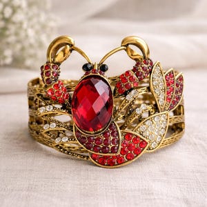 May include: A gold-toned cuff bracelet featuring a lobster design. The bracelet is adorned with red and clear crystals, including a large, faceted red stone at the center. The lobster's claws and antennae are detailed.