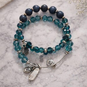May include: Two bracelets with blue beads and silver charms. One bracelet features dark blue, round beads. The other bracelet has faceted, teal-colored beads. Silver charms include a heart and a tag.