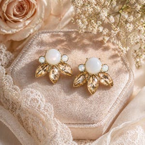 May include: A pair of gold-toned stud earrings. Each earring features a large, round, white pearl-like center stone, surrounded by smaller, opalescent stones and amber-colored, petal-shaped crystals. The earrings are displayed on a velvet surface.