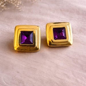 May include: A pair of square gold-toned earrings with a deep purple square gemstone center. The earrings have a layered design, with the gemstone set within a square frame. The background is a soft, light pink fabric.