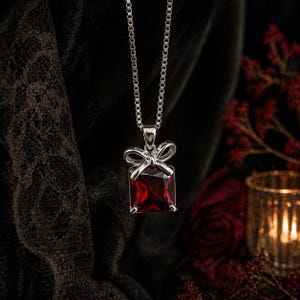 May include: A silver necklace with a rectangular, deep red gemstone pendant. The pendant is framed by a silver bow design. The chain is a delicate box chain. The background is dark, with hints of red and gold.