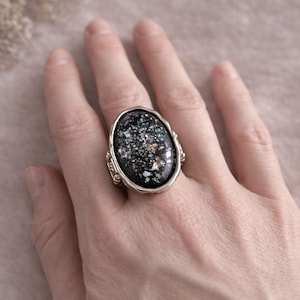 May include: Oval-shaped silver ring with a large, dark stone. The stone has a speckled pattern of white, gray, and teal. The ring's band has decorative detailing on the sides. A unique piece of jewelry.