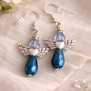 May include: A pair of angel-themed earrings. Each earring features a silver-toned wing design, a light blue bead, and a teardrop-shaped, faceted, dark blue bead. The earrings hang from silver-colored ear wires.