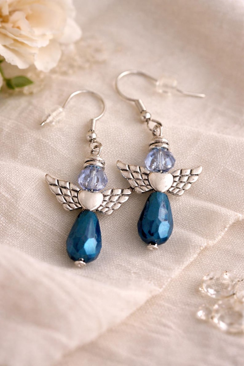 May include: A pair of angel-themed earrings. Each earring features a silver-toned wing design, a light blue bead, and a teardrop-shaped, faceted, dark blue bead. The earrings hang from silver-colored ear wires.