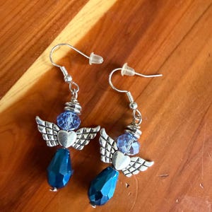 May include: A pair of dangle earrings featuring an angel design. Each earring has silver-toned wings, a heart, a blue bead, and a faceted blue teardrop-shaped bead. The earrings hang from silver-colored ear wires.