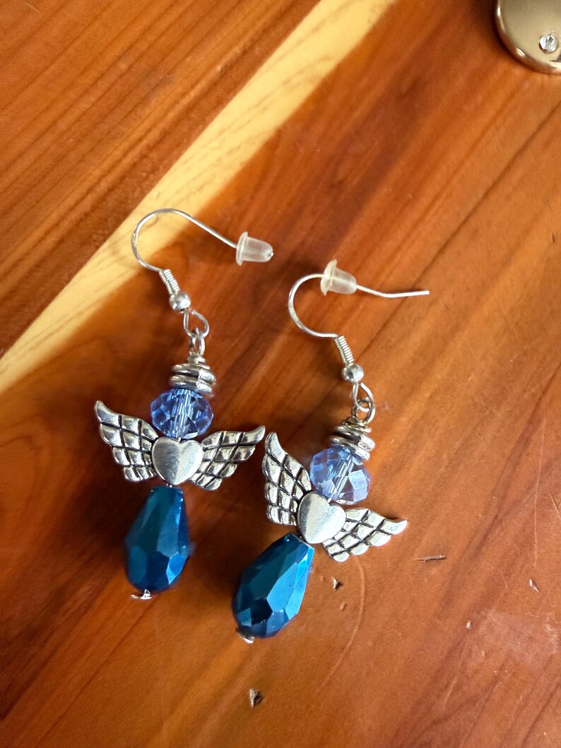 May include: A pair of dangle earrings featuring an angel design. Each earring has silver-toned wings, a heart, a blue bead, and a faceted blue teardrop-shaped bead. The earrings hang from silver-colored ear wires.