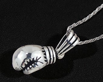 May include: Silver-colored boxing glove pendant necklace. The pendant is detailed with stitching and a starburst design. The necklace chain is a thin, twisted rope style. The background is a solid black.