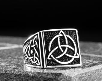 May include: A silver-colored ring with a square face featuring a Celtic knot design. The ring is set on a gray, textured surface against a dark background. The Celtic knot is a symbol of interconnectedness.