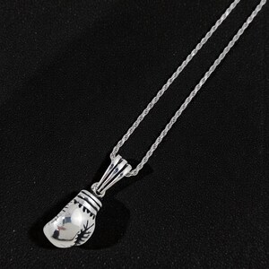 May include: A silver boxing glove pendant necklace. The pendant is detailed with stitching and a rope chain. The necklace is displayed against a black background.
