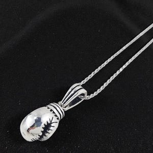 May include: A silver-colored pendant necklace featuring a boxing glove design. The pendant has intricate detailing, including stitching and a ribbed cuff. The necklace chain is a delicate, twisted rope style, and the background is a dark fabric.