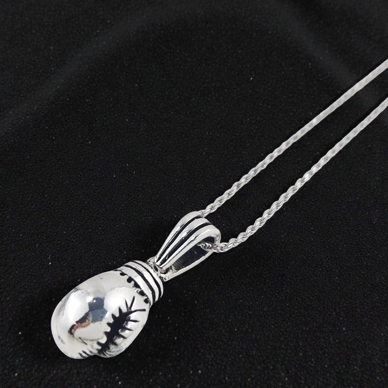 May include: A silver-colored pendant necklace featuring a boxing glove design. The pendant has intricate detailing, including stitching and a ribbed cuff. The necklace chain is a delicate, twisted rope style, and the background is a dark fabric.