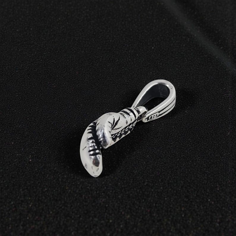 May include: Silver boxing glove pendant with intricate detailing. The pendant features a loop for a chain and is designed to resemble a miniature boxing glove. The metal has a polished finish, reflecting light.