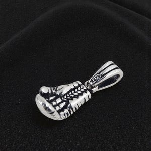 May include: A silver pendant in the shape of a boxing glove. The pendant is detailed with textured lines and a loop for a chain. The metal has a shiny finish. The background is a dark, solid color.