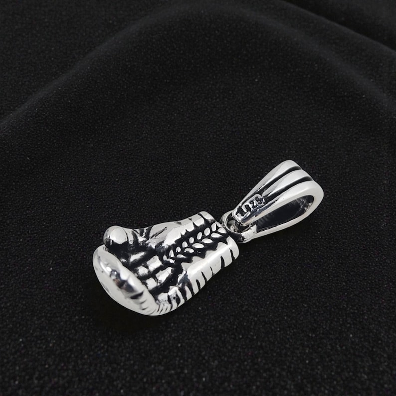 May include: A silver pendant in the shape of a boxing glove. The pendant is detailed with textured lines and a loop for a chain. The metal has a shiny finish. The background is a dark, solid color.