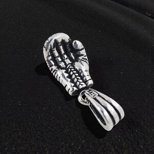 May include: A silver-colored pendant in the shape of a boxing glove. The glove is detailed with lines and textures, and is connected to a separate loop for a chain. The background is a dark, textured surface.