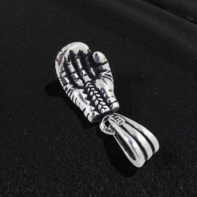 May include: A silver-colored pendant in the shape of a boxing glove. The glove is detailed with lines and textures, and is connected to a separate loop for a chain. The background is a dark, textured surface.