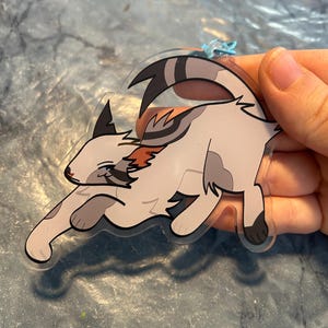 Op de afbeelding: Een heldere acryl kattenbedel in een speelse pose. De kat heeft een wit lichaam met grijze en oranje markeringen, zwarte strepen op de staart en een blauwe lus om op te hangen. De bedel wordt door een hand vastgehouden.