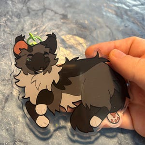 Pu&ograve; includere: Un ciondolo in acrilico trasparente raffigurante un gatto dei cartoni animati con una coda e zampe soffici. Il gatto ha un mantello grigio e nero con accenti marroni e un anello portachiavi verde.