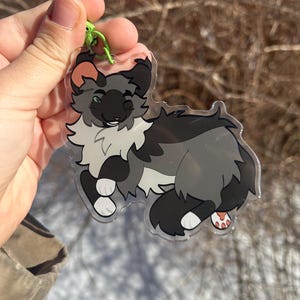 Pu&ograve; includere: Un portachiavi in acrilico trasparente con un gatto dei cartoni animati con pelliccia grigia, nera e bianca. Il gatto ha occhi verdi e cuscinetti rosa. Un fermaglio verde &egrave; attaccato alla parte superiore del portachiavi.